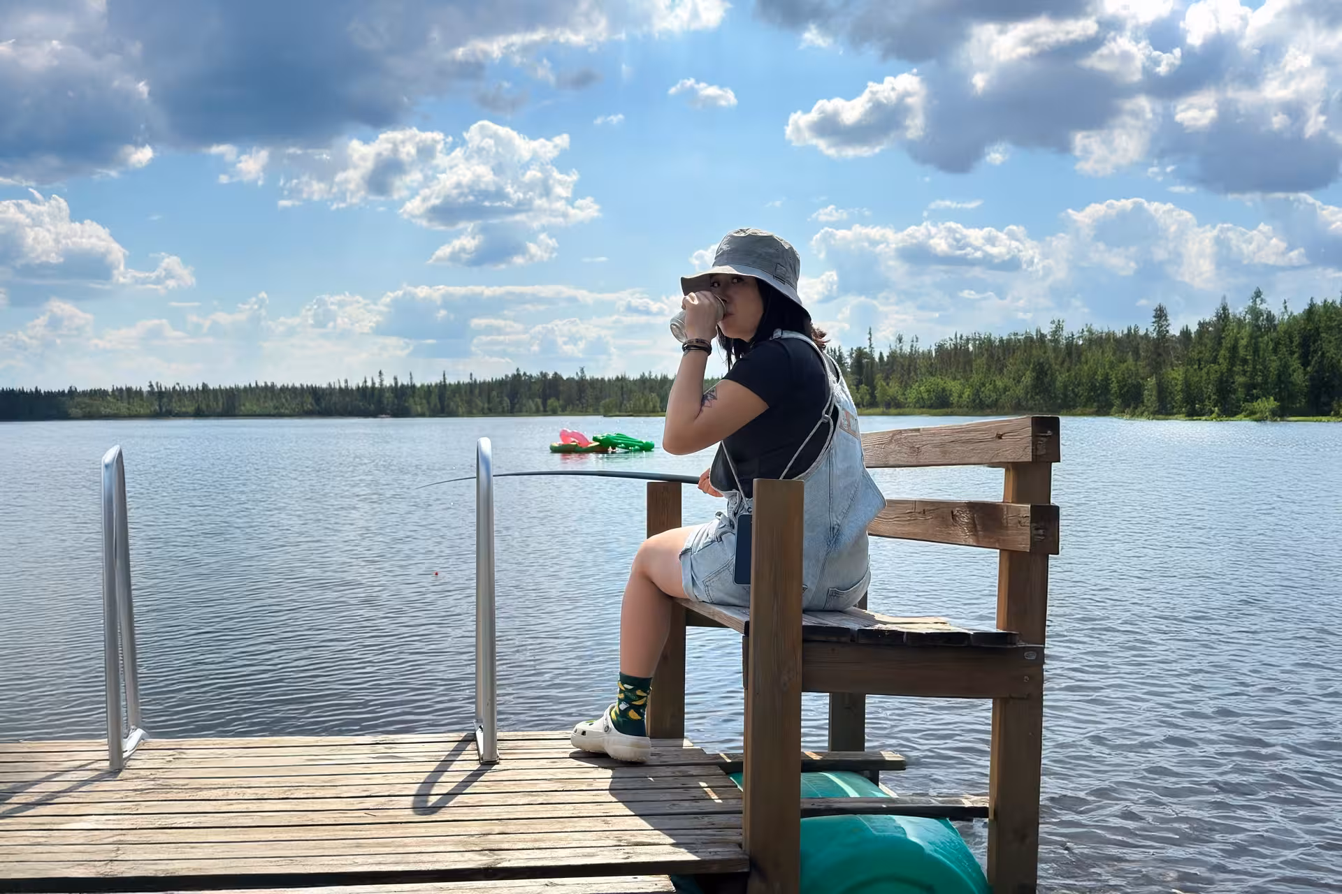 Person enjoying a drink on a wooden dock by a tranquil Arctic lake under a clear blue sky in Rovaniemi.