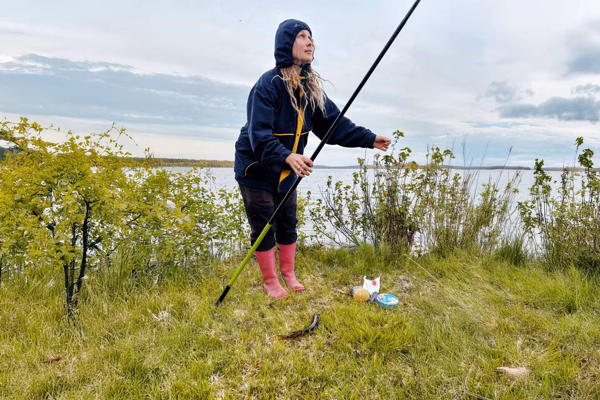 Fisher in raincoat and boots holding a fishing rod by an Arctic lakeside in Rovaniemi, surrounded by greenery.