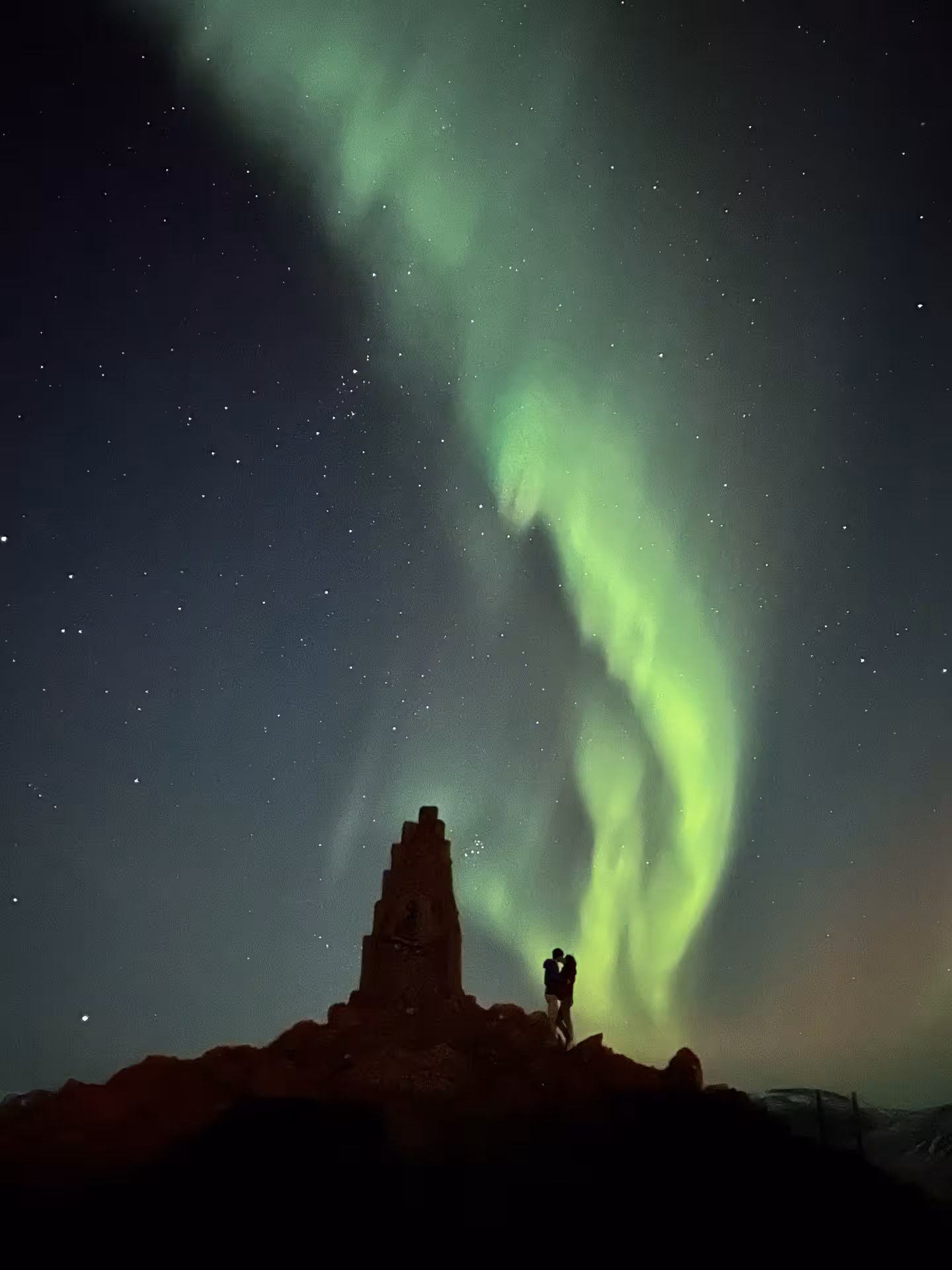 Couple silhouetted on rocky ridge beneath sweeping green aurora, private Arctic Odyssey husky and aurora tour