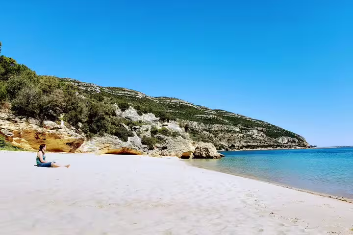 Person relaxing on Arrábida's pristine beach with clear blue waters and scenic cliffs, perfect for snorkeling and wine tours.