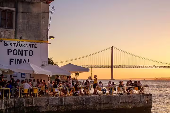 Seaside dining at sunset near a bridge in Lisbon, showcasing the scenic beauty of Arrábida & Sesimbra on a wine tasting day trip.