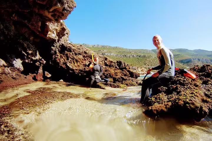 Two snorkelers explore rocky coastal waters during an Arrábida adventure, with clear skies and lush hills in the background.