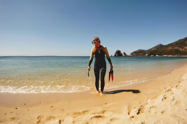 A snorkeler in a wetsuit walks along the pristine beach of Arrábida with clear blue waters, ready for an adventure from Lisbon.