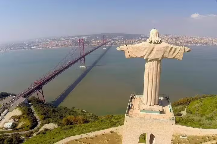 Aerial view of Lisbon's iconic Christ the King statue overlooking the 25th of April Bridge, a highlight on Arrábida/Setúbal private tours.