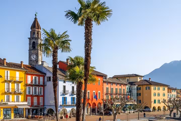 Colorful lakeside promenade in Ascona with palm trees and historic architecture against mountain backdrop.