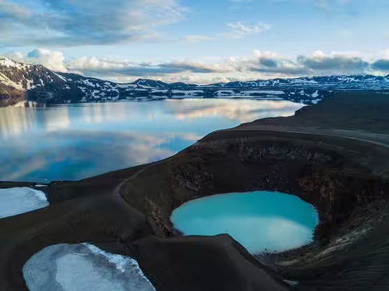 Aerial view of Askja caldera with vivid blue crater lake surrounded by dark volcanic terrain and snow-capped mountains
