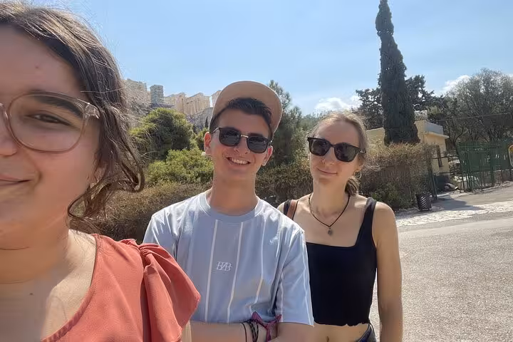 Three young tourists smiling during a self-guided walking tour in Athens with the Acropolis visible in the background
