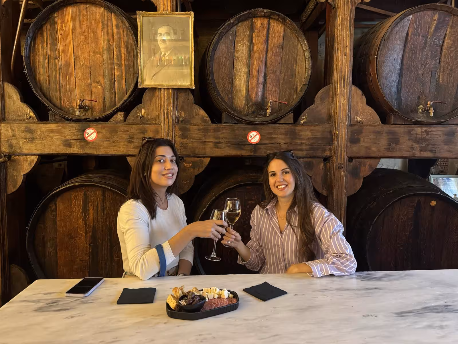 Couple clinking glasses in a rustic Athens wine bar with oak barrels, sampling Greek wine and meze on evening tour