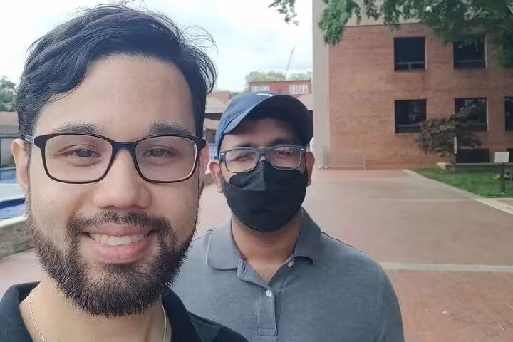 Two men taking a selfie outside a brick building, one wearing glasses and beard, other in cap and mask