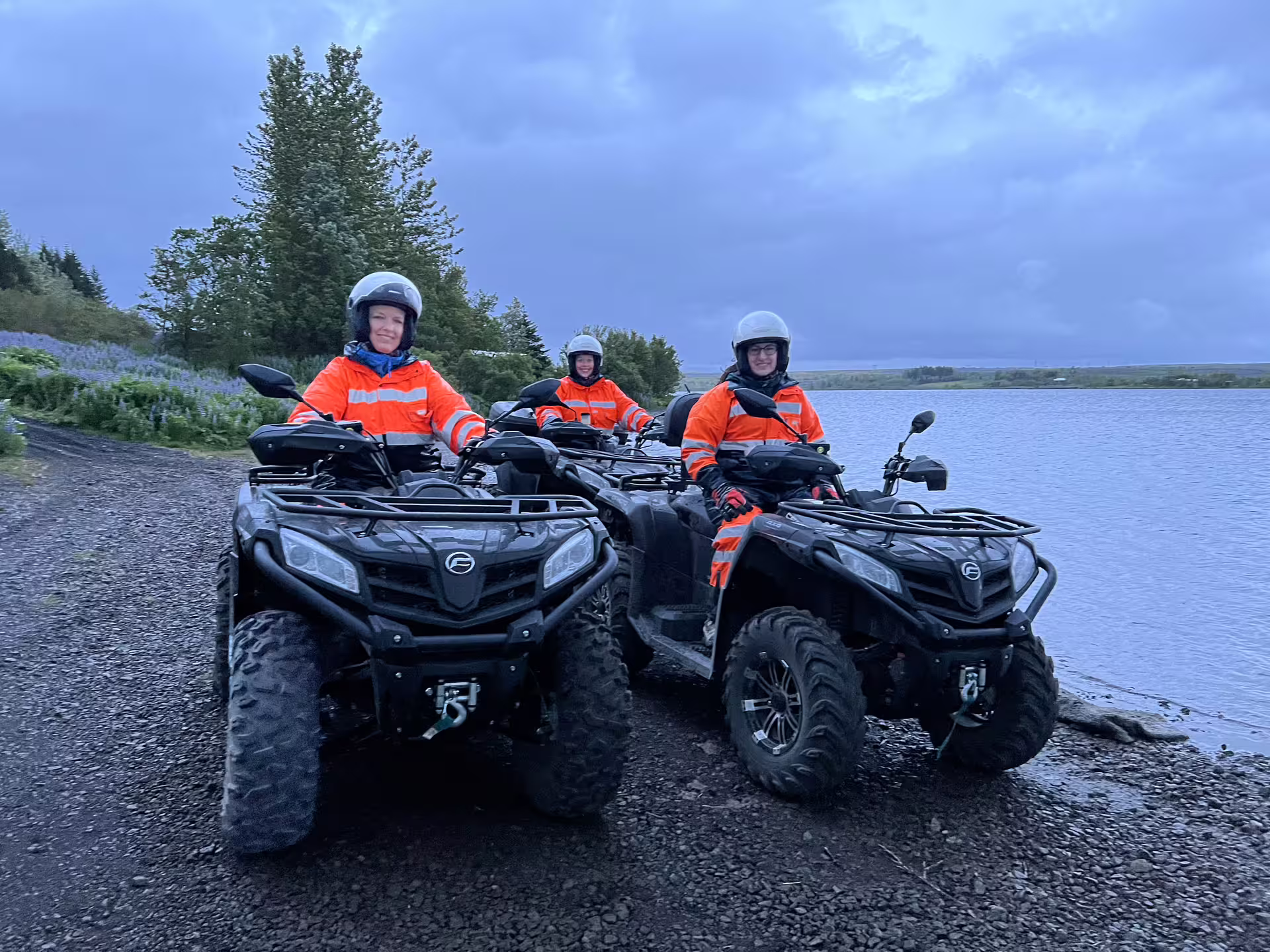 Three riders in orange safety gear on black ATVs beside a lake in Reykjavik under dramatic cloudy skies