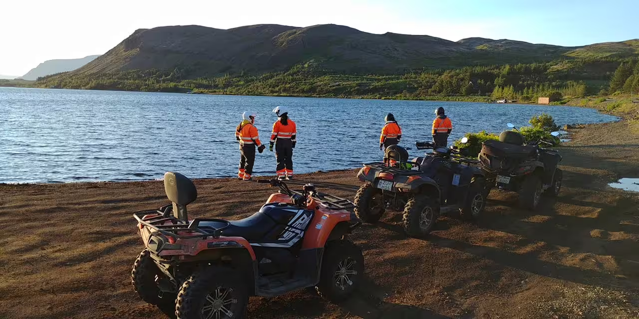 Group of four people in orange safety gear standing by a scenic lake with ATVs parked on shore near Reykjavik