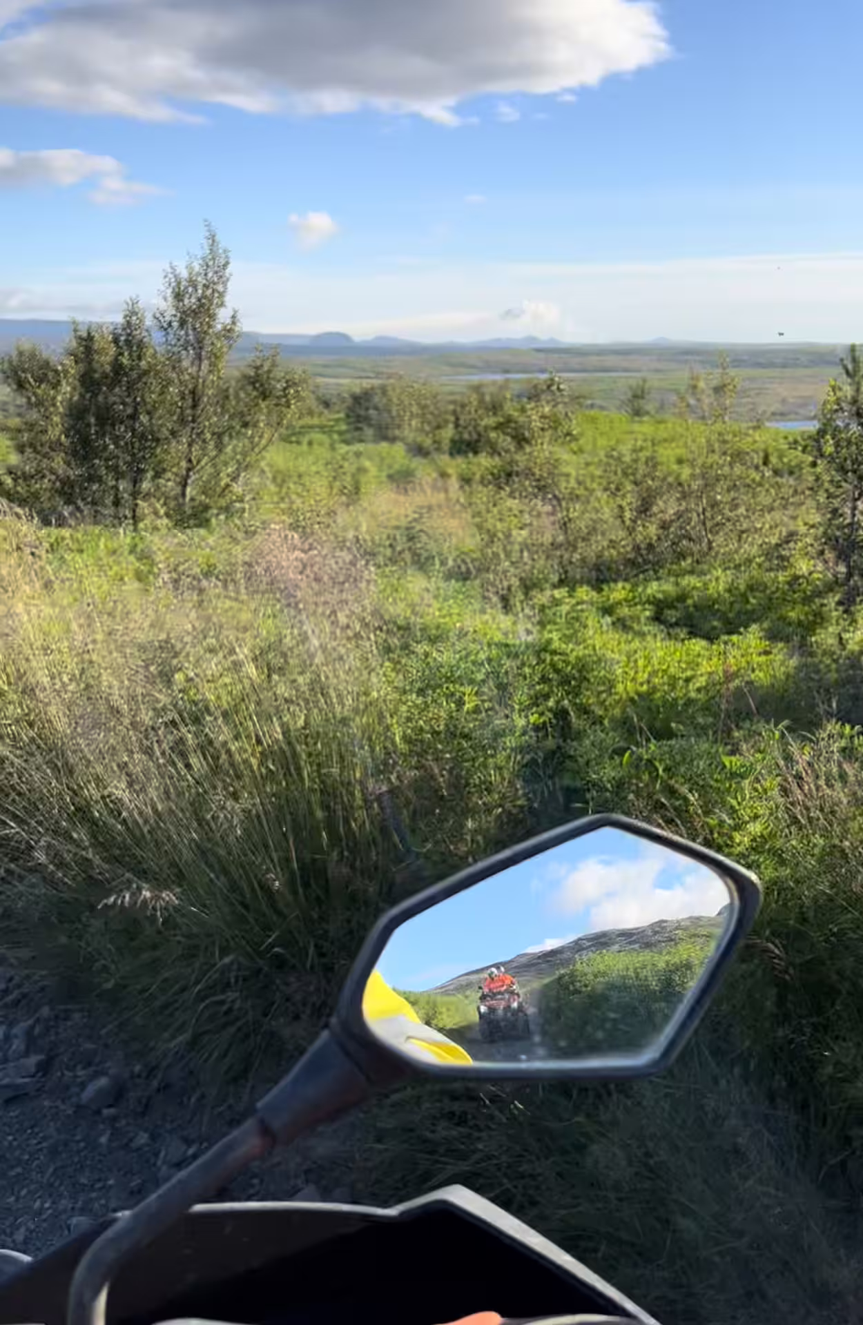 ATV side mirror reflecting another quad bike rider on scenic Iceland trail with green landscape and blue sky