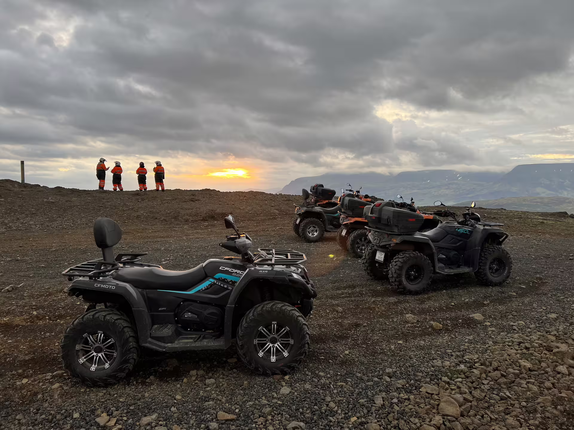 CFMOTO ATVs parked on rocky terrain with riders in orange gear watching sunset over Icelandic mountains