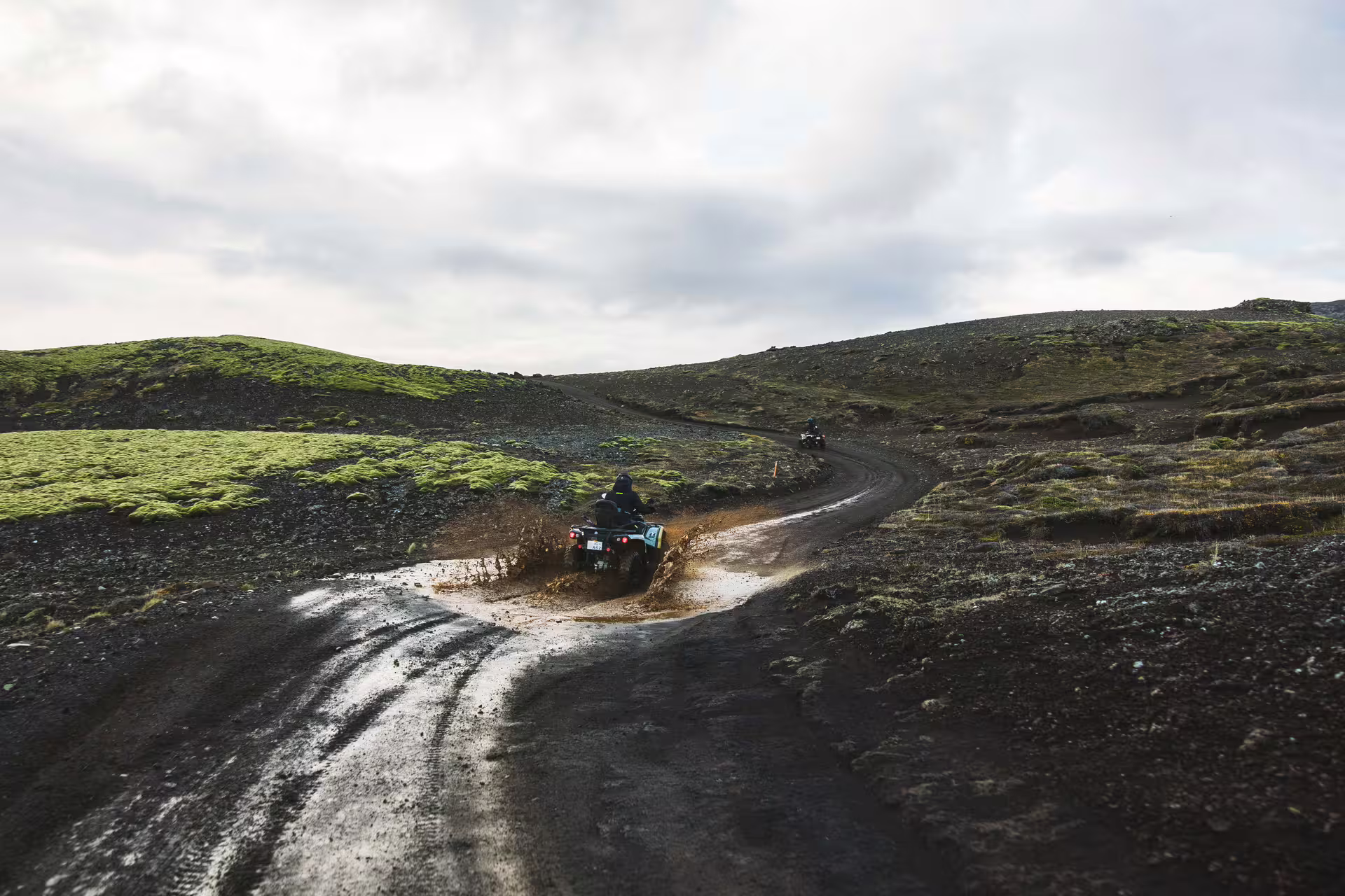 ATV riders splashing through muddy puddle on black lava trail with green moss hills in Iceland
