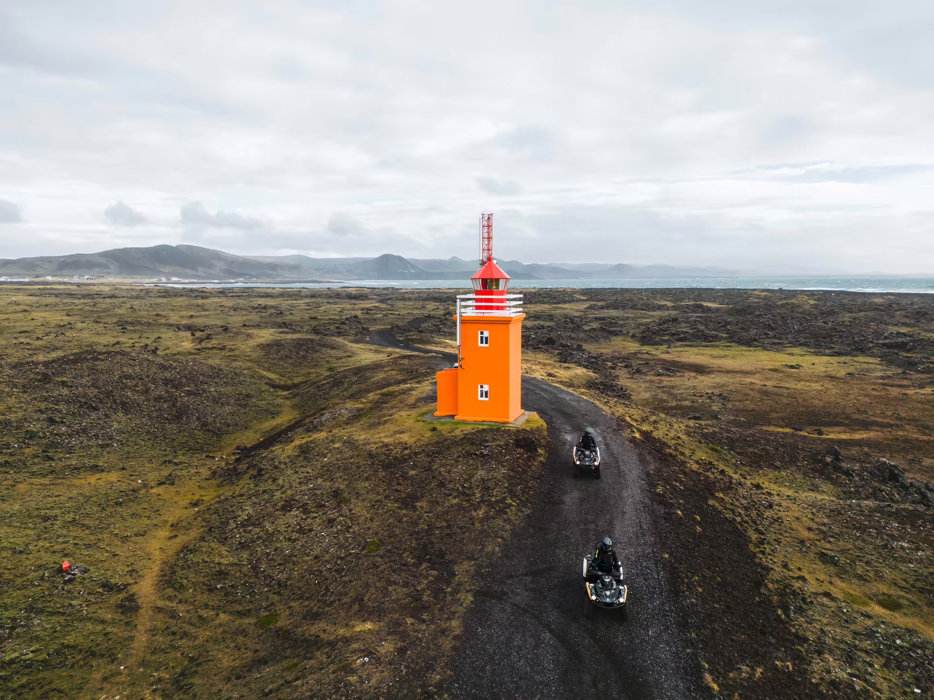 Aerial view of two ATVs riding a gravel trail toward an orange lighthouse on Iceland's lava fields