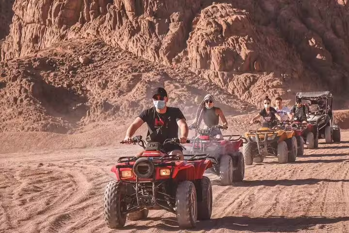 Group riding ATV quad bikes through Marsa Alam desert mountains on safari tour with camel ride and Bedouin dinner