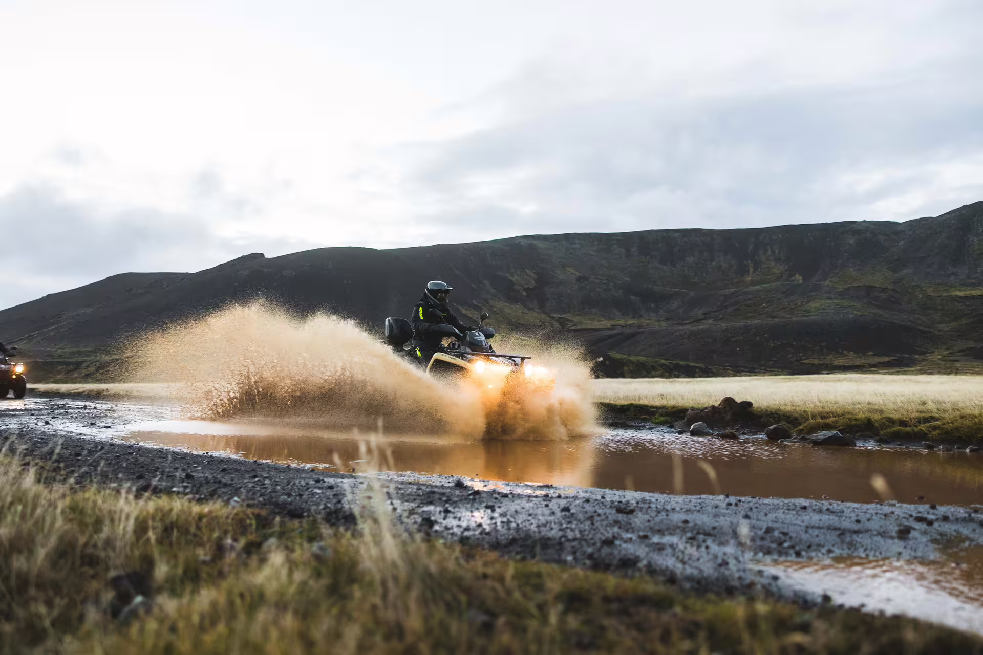 ATV rider splashing through muddy water on Iceland lava trail with dark volcanic mountains in background