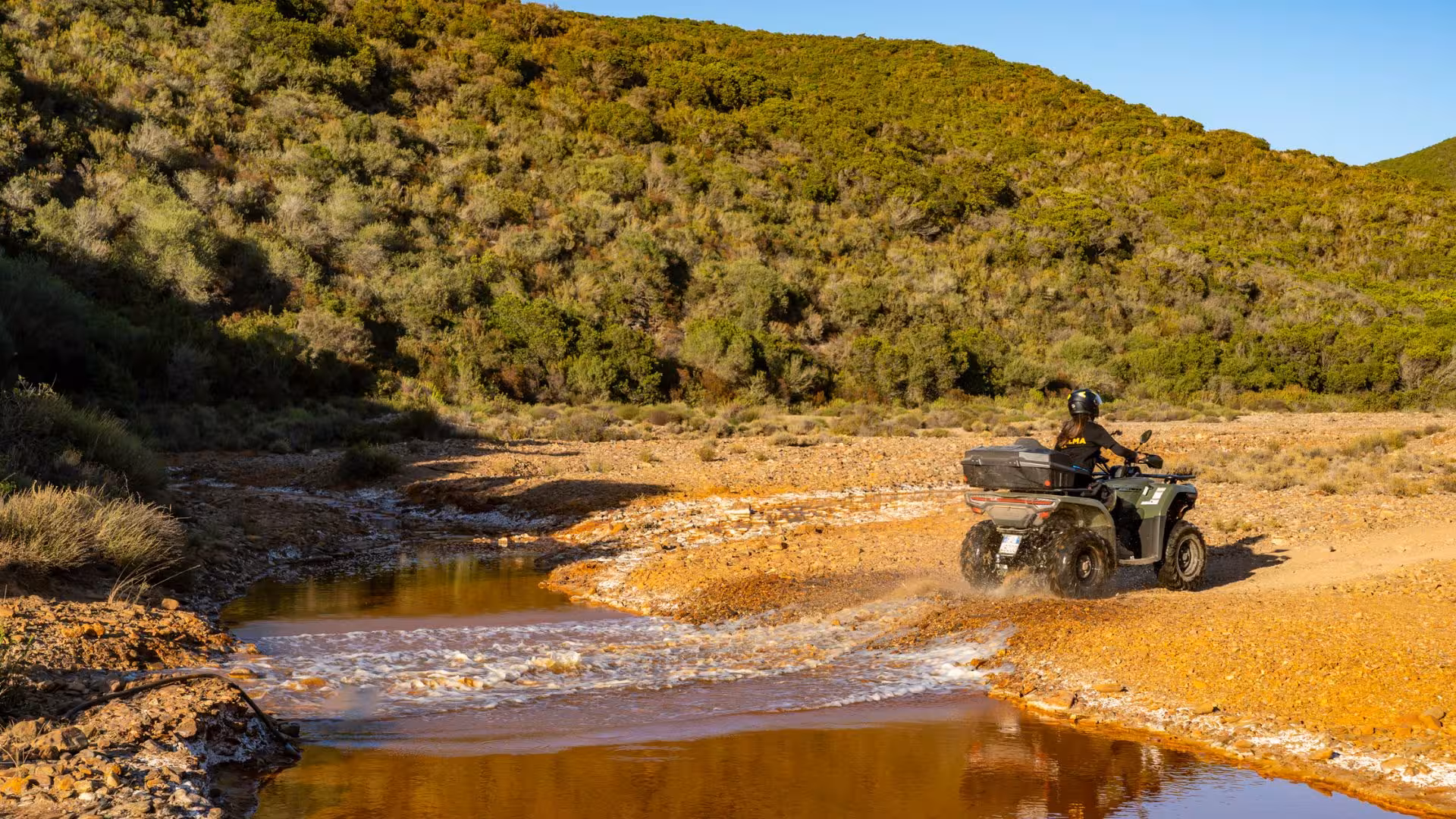 ATV rider crossing a scenic stream on rugged Arbus terrain during an adventurous excursion.