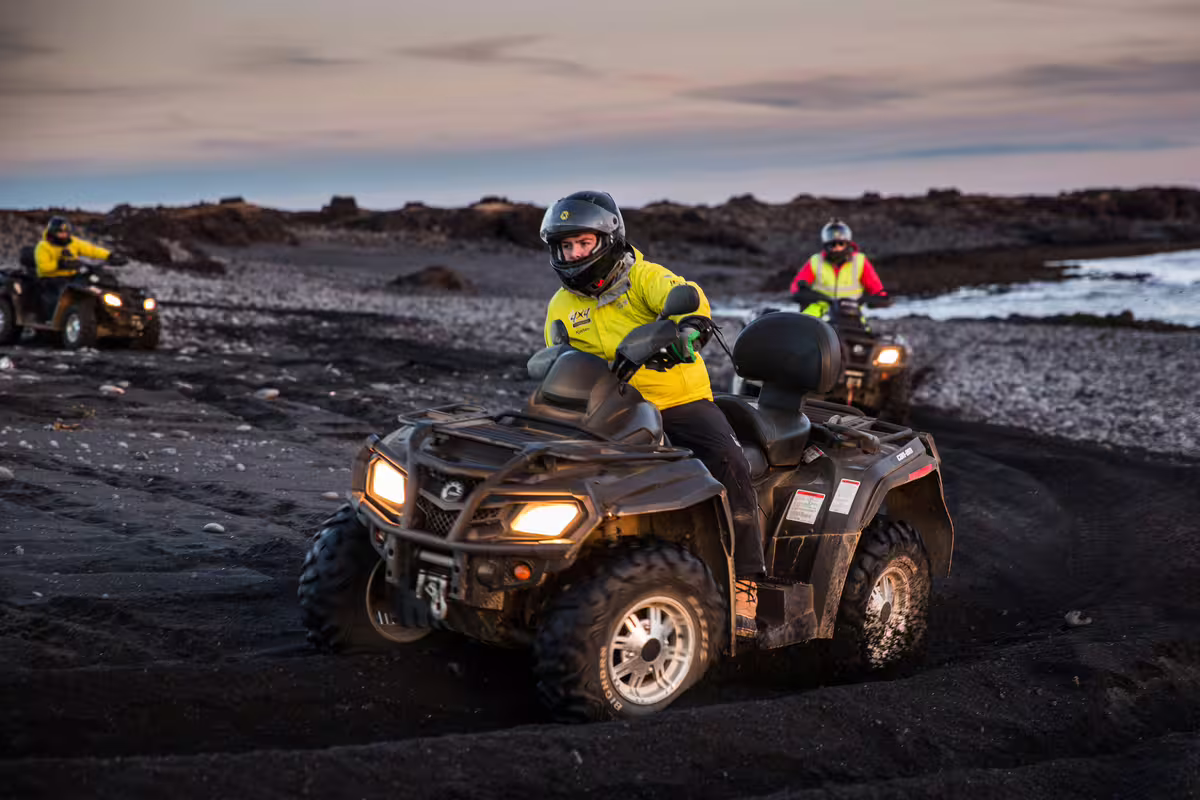 Riders on ATVs wearing yellow helmets and jackets crossing Iceland's black sand beach at dusk