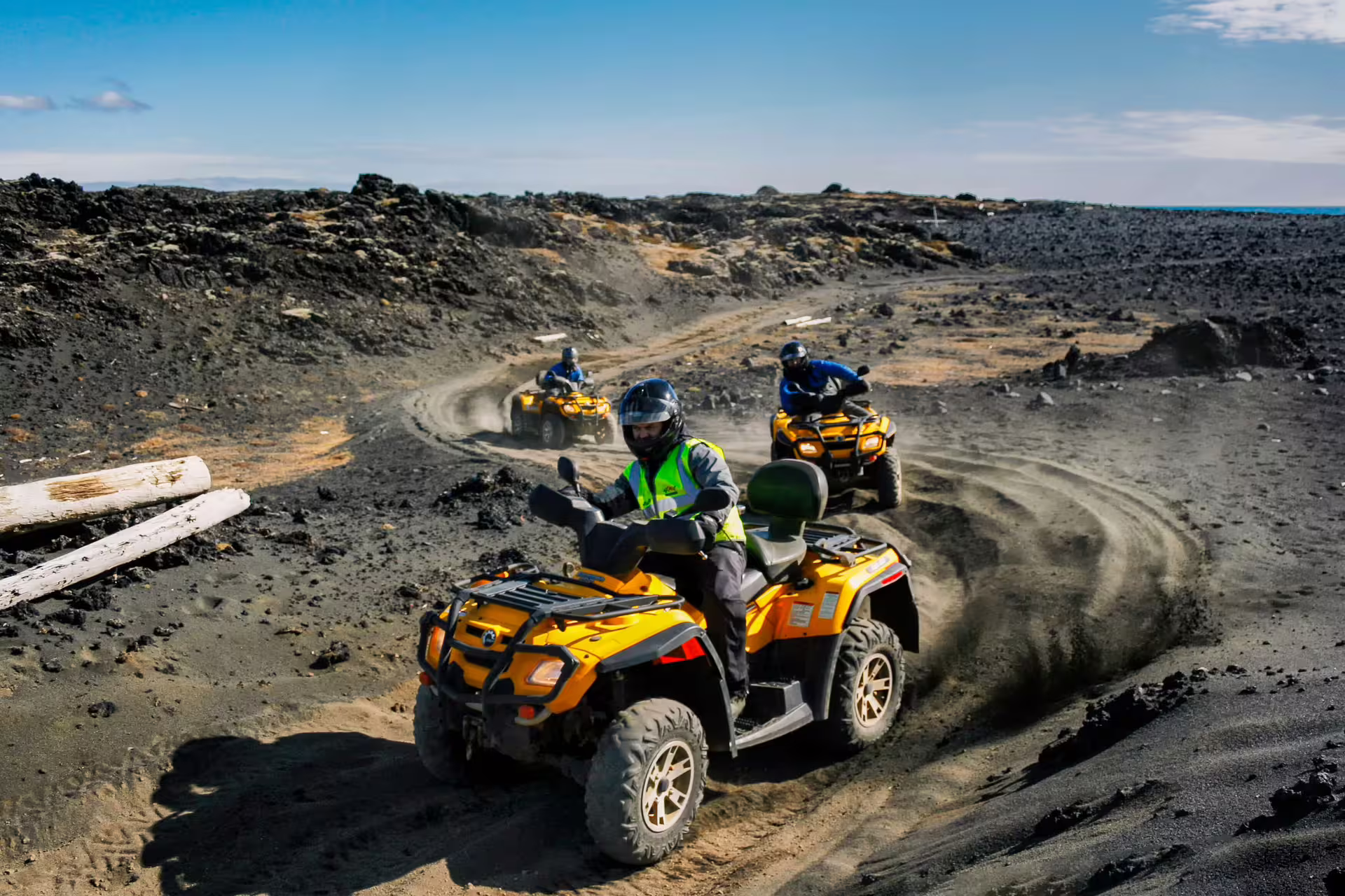 Three riders on yellow ATVs navigating black lava sand trails in Iceland under blue sky