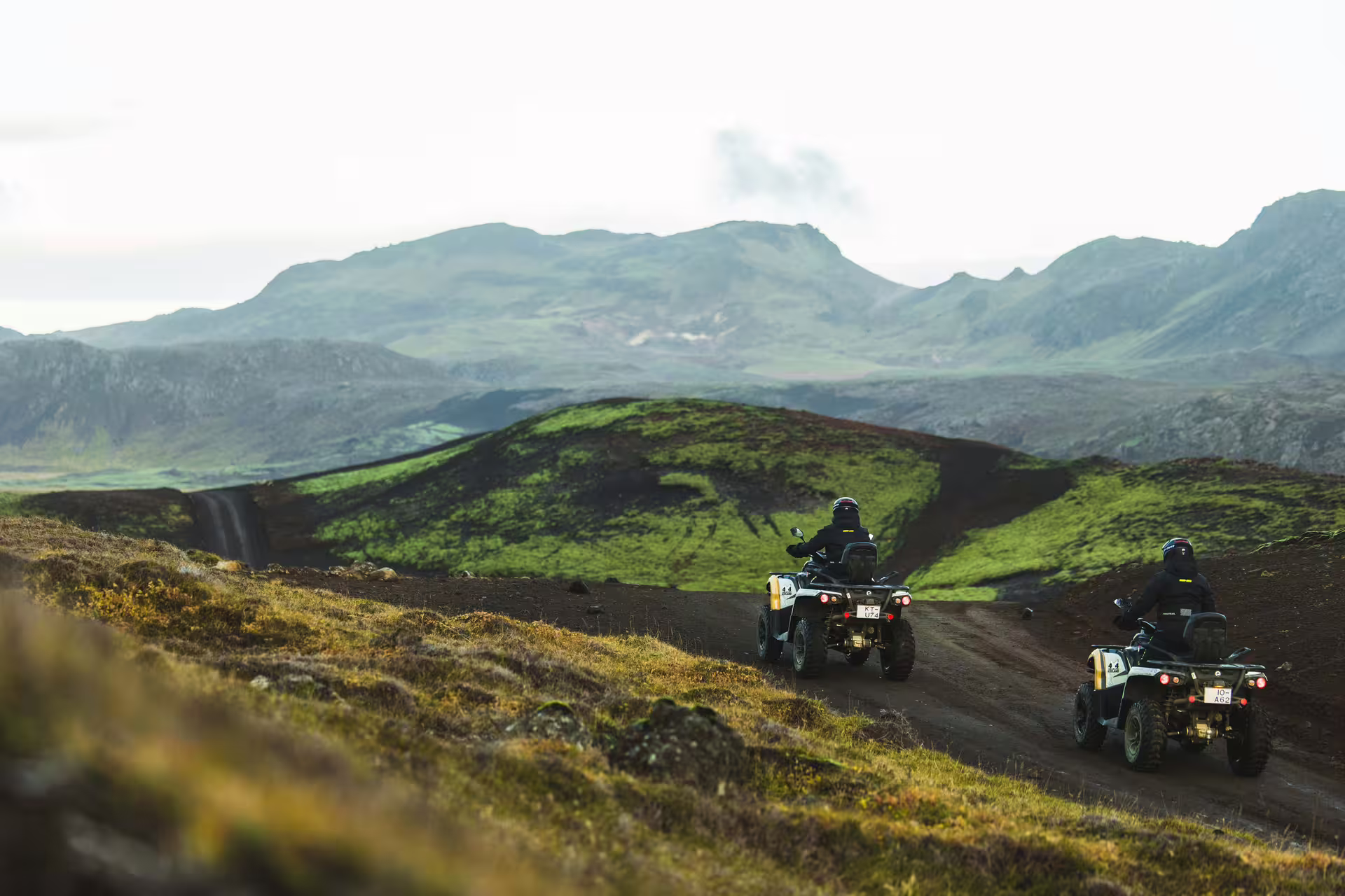 Two ATV riders on black lava trail with green volcanic mountains in Iceland background
