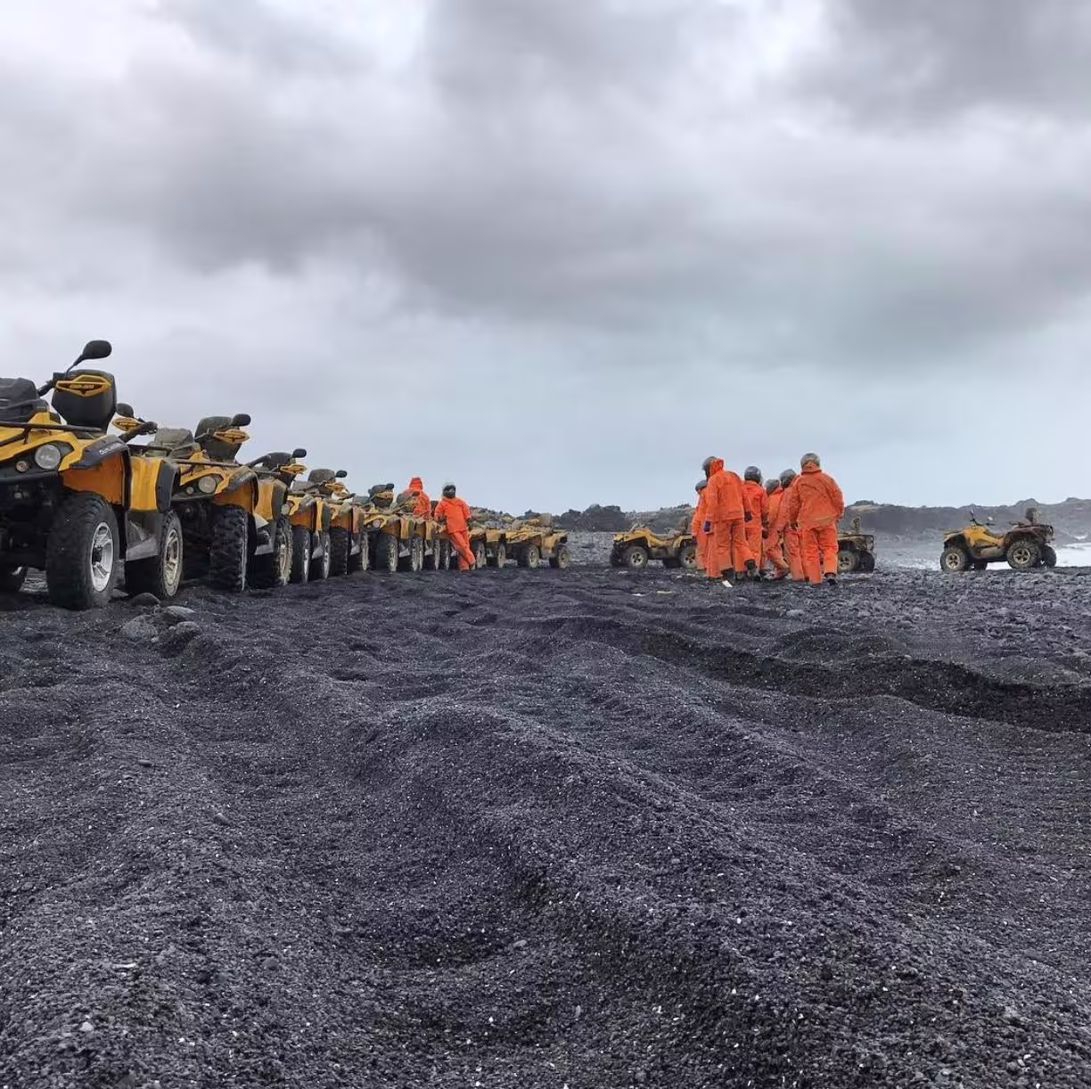 Group in orange suits with yellow ATVs lined up on black sand beach in Iceland under cloudy skies