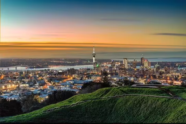 Stunning sunset over Auckland cityscape with Sky Tower, viewed from a hilltop, highlighting vibrant urban lights.