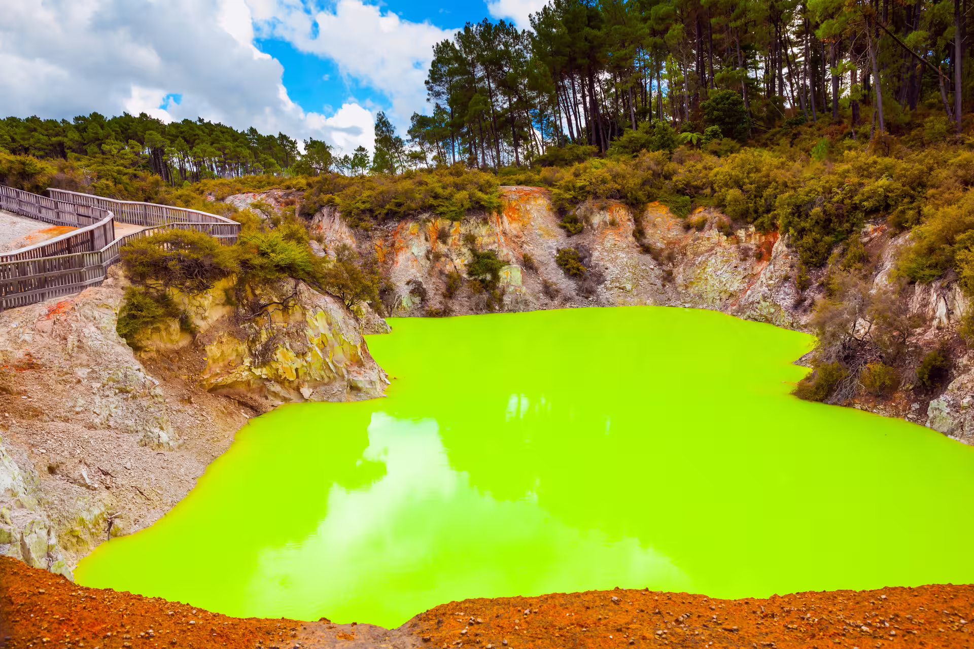 Vibrant green geothermal lake surrounded by rocky cliffs and forested areas in Rotorua's thermal landscape.