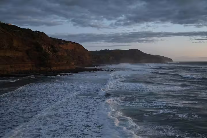 Waves crashing against Auckland's dramatic cliffs at sunset, perfect for a romantic tour with winery and thermal pool experience.