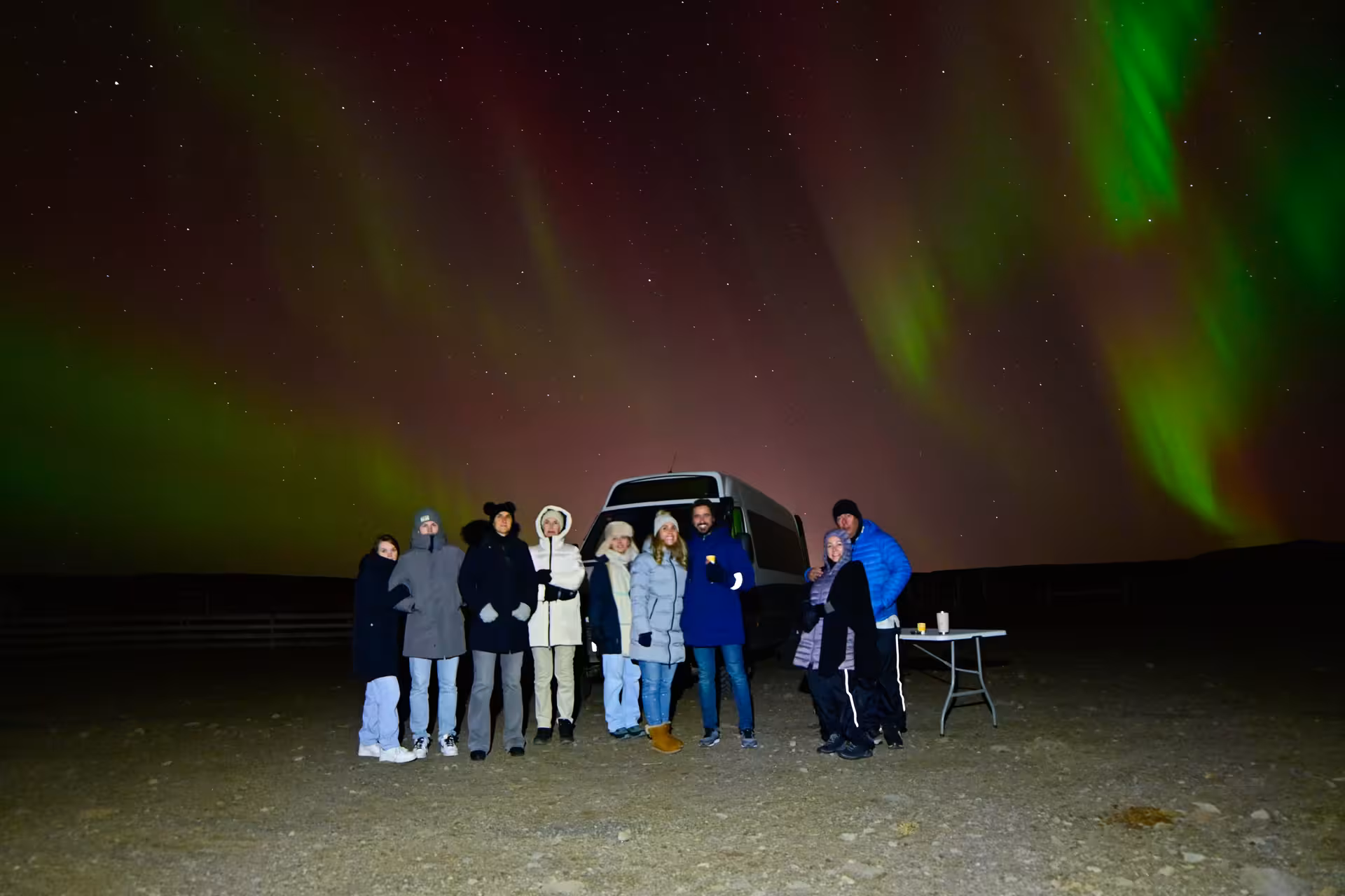 Tour group posing before colorful aurora borealis with red and green lights beside a 4x4 van on Iceland northern lights tour