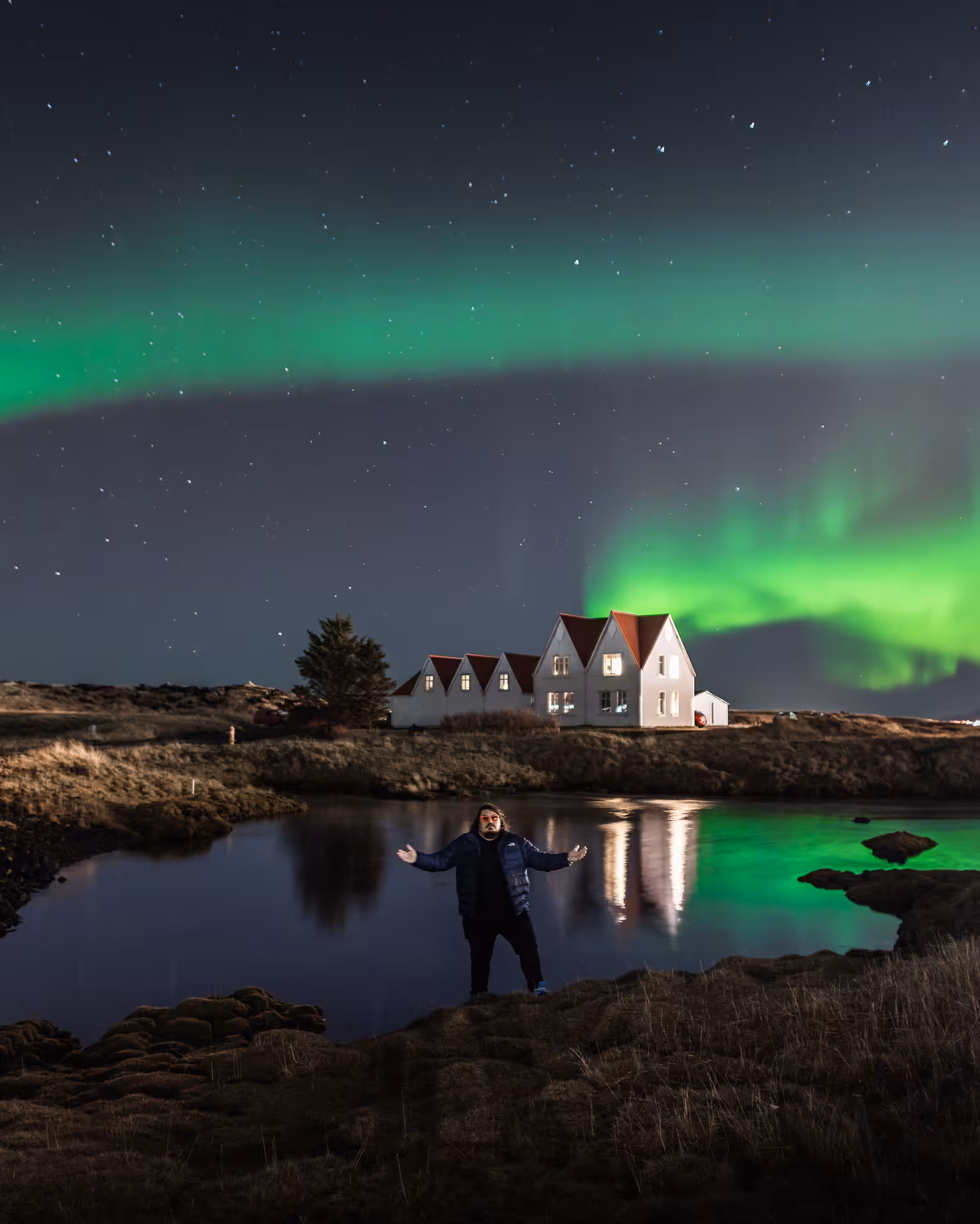 Man standing by a reflective lake beneath vivid green aurora borealis over Icelandic white houses at night
