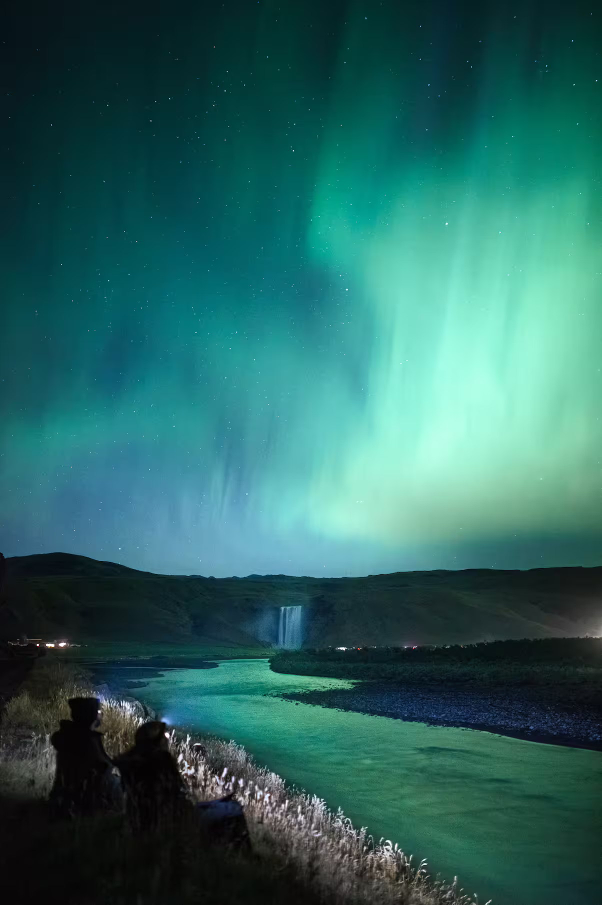 Photographer and guests watching vibrant green aurora borealis over an Iceland river and waterfall at night