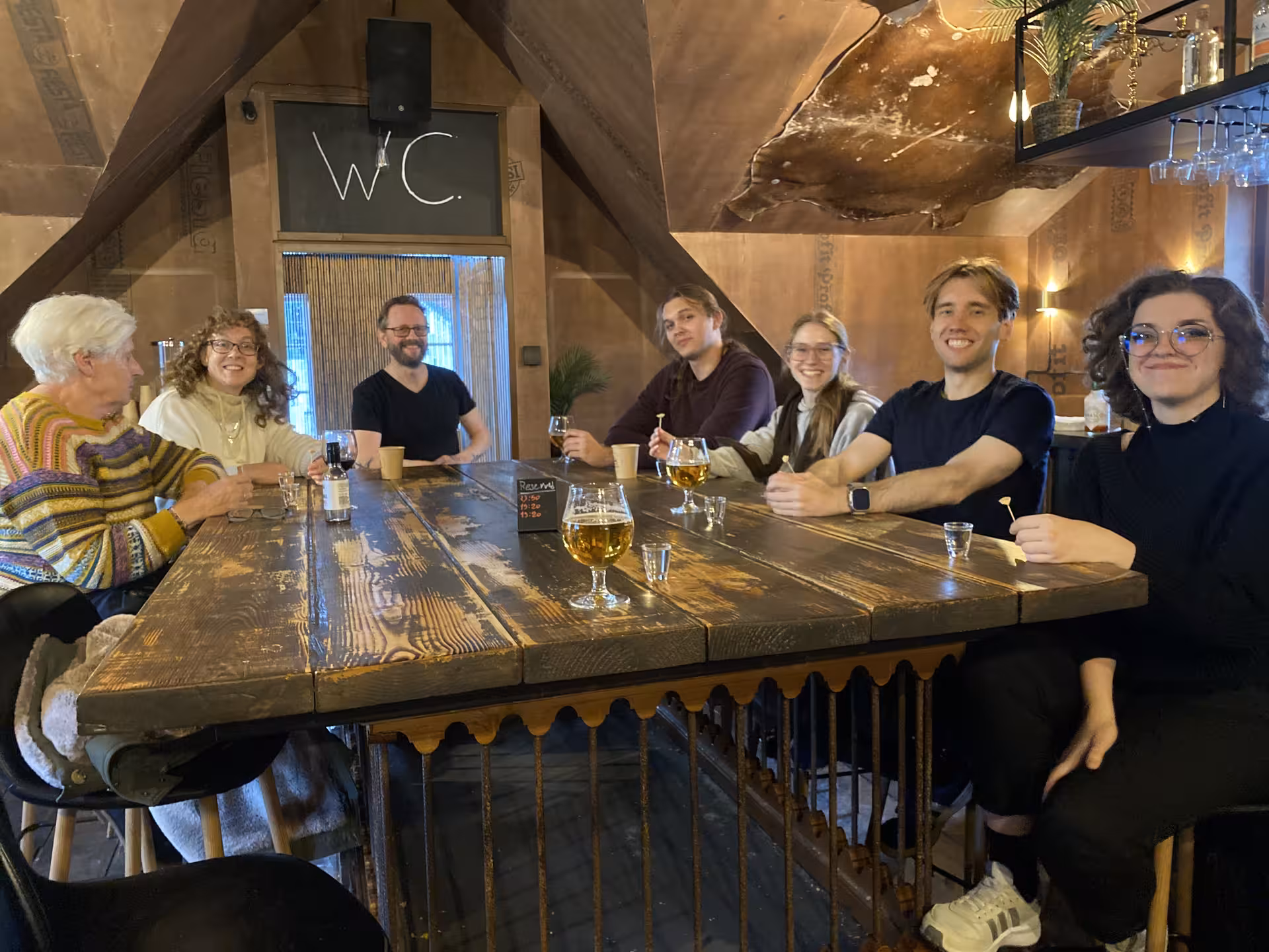 Group of seven people smiling around a rustic wooden table with beer glasses in a cozy Icelandic bar
