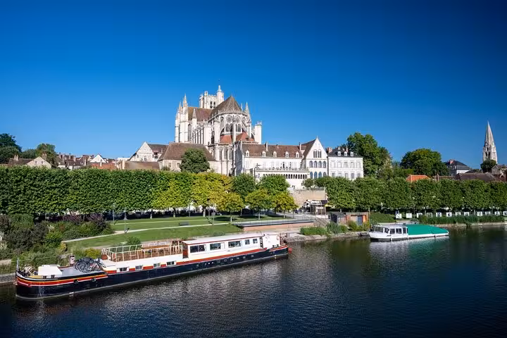 Auxerre Cathedral rising above the Yonne River with moored barges and lush green trees on a sunny day