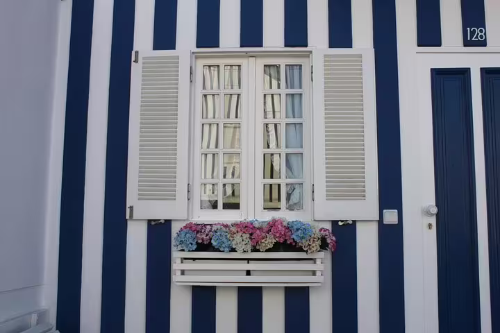 Charming striped house facade with a white window and colorful flowers in Costa Nova, Aveiro tour highlight.