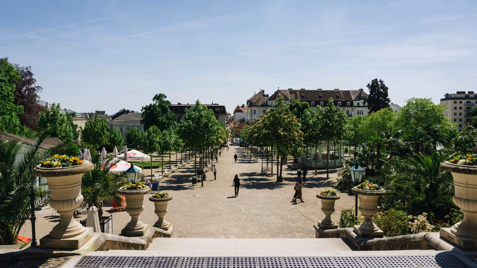 Tree-lined promenade in Baden-Baden park with stone urns, flower planters, and historic buildings in background
