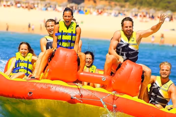 Group of friends laughing on a fast banana boat slider ride, splashing through clear blue sea near a sunny Mediterranean beach