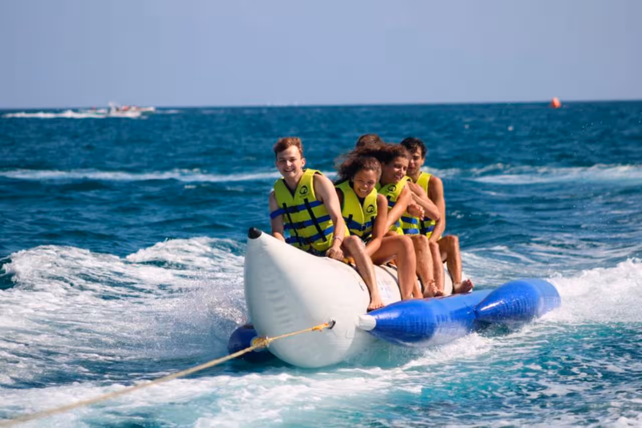 Group of friends enjoying a thrilling banana slider ride on turquoise sea waves, wearing life vests on a summer adventure