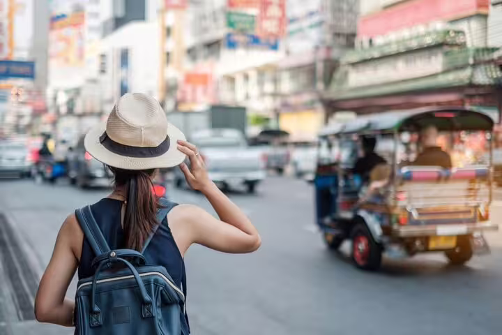 Female tourist with backpack and hat hailing a tuk-tuk on a busy Bangkok street