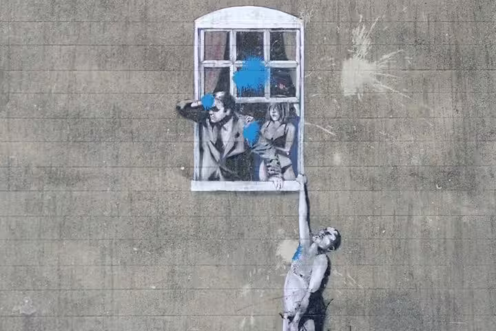 Banksy street art mural in Bristol showing man hanging from window ledge with couple inside