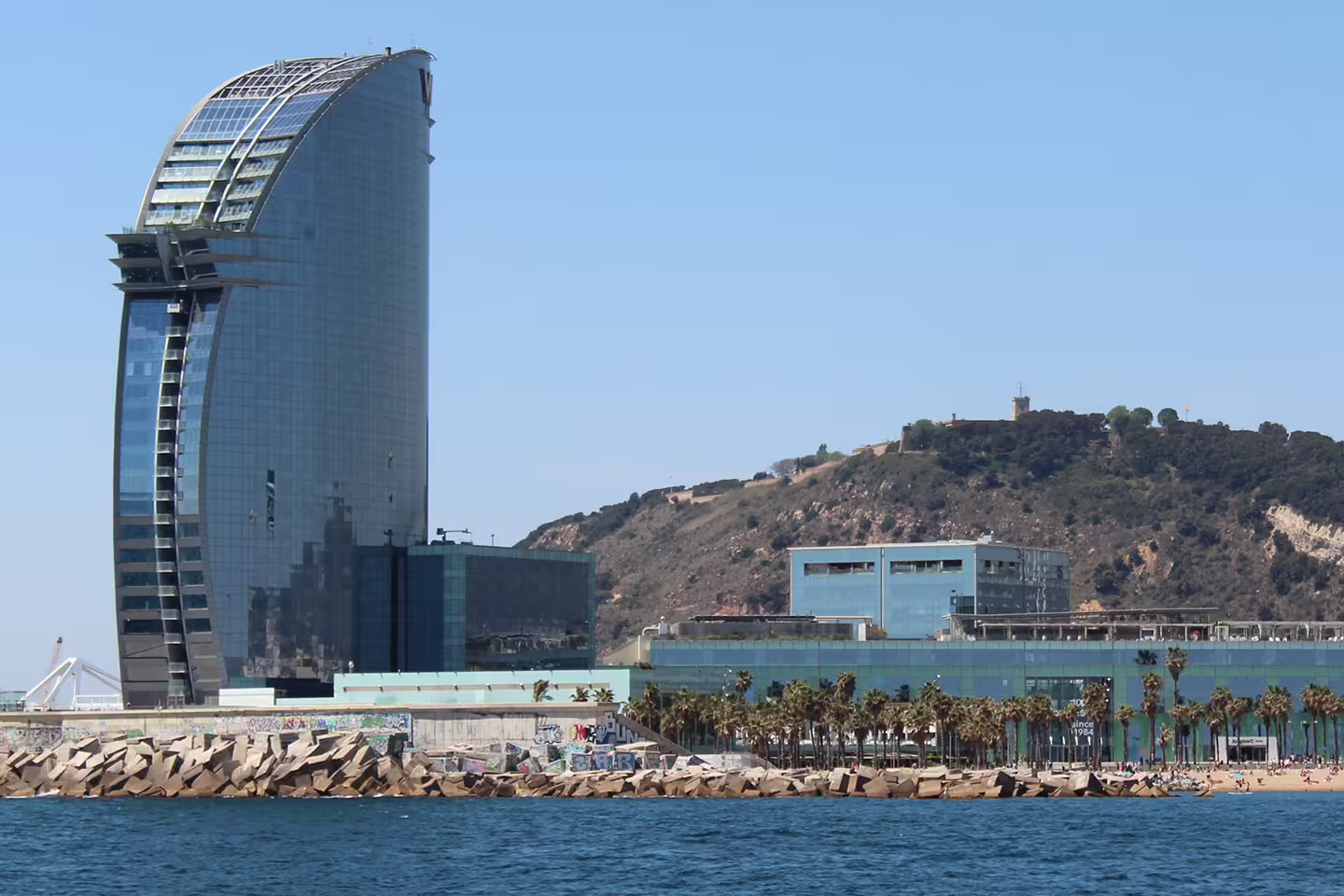 Iconic sail-shaped building by Barcelona's coastline with a scenic view of hills in the background.