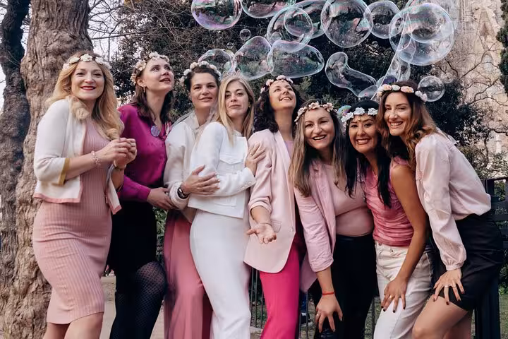 Friends in pink outfits and flower crowns pose under bubbles in a leafy Barcelona park, capturing a joyful photoshoot moment.