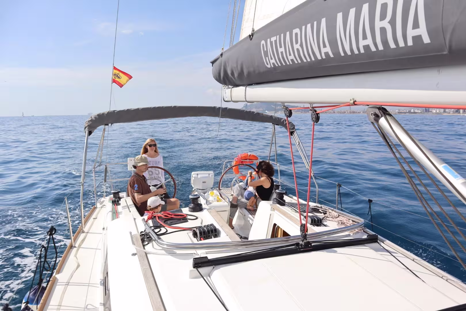Sailing experience in Barcelona with people enjoying a sunny day on a boat, featuring the Spanish flag and open sea views.