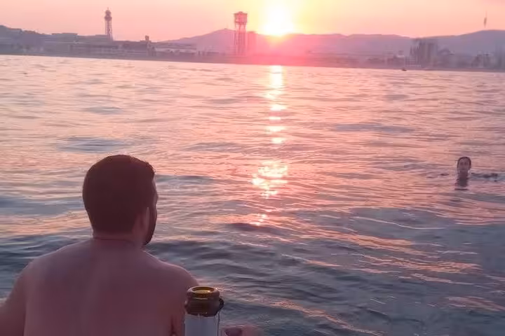 Guests relax on a sailboat at Barcelona sunset with open bar drinks, city skyline glowing over the sea