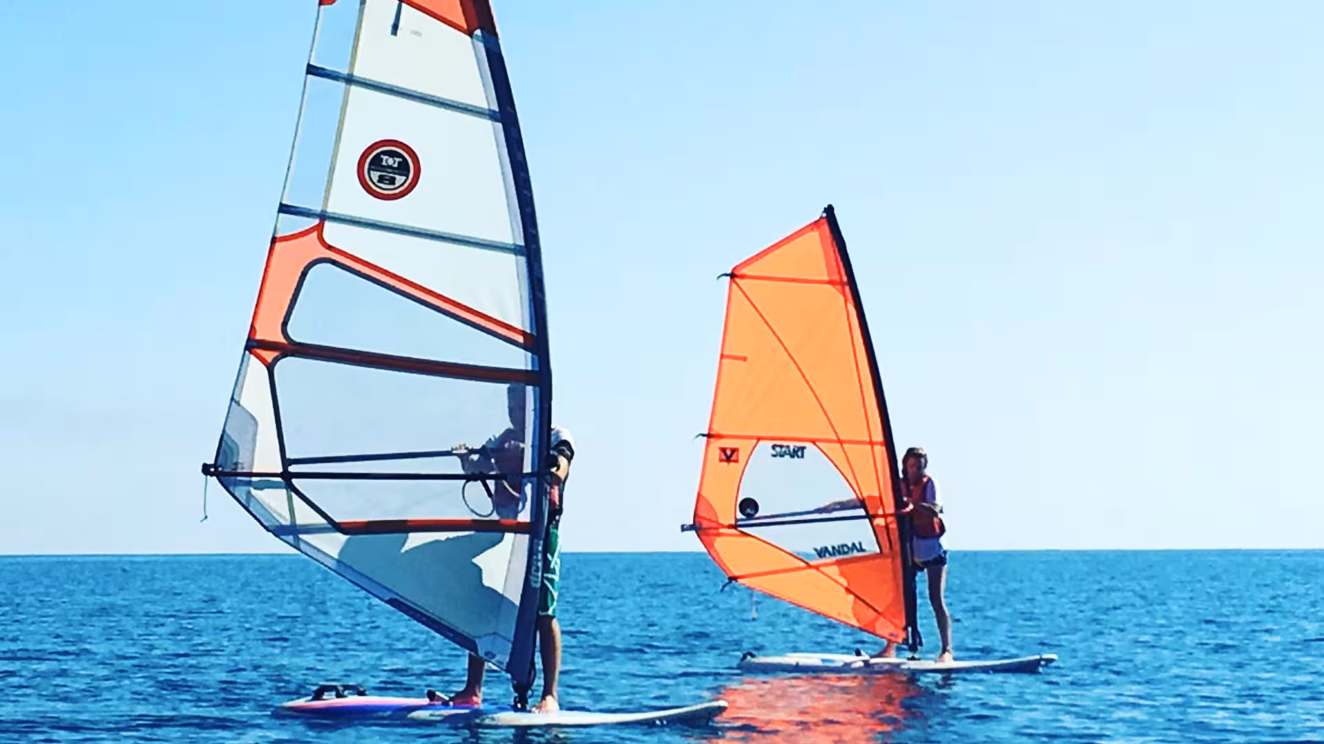 Two windsurfers balancing on calm waters in Bari Sardo, ideal for beginners learning the basics of windsurfing.