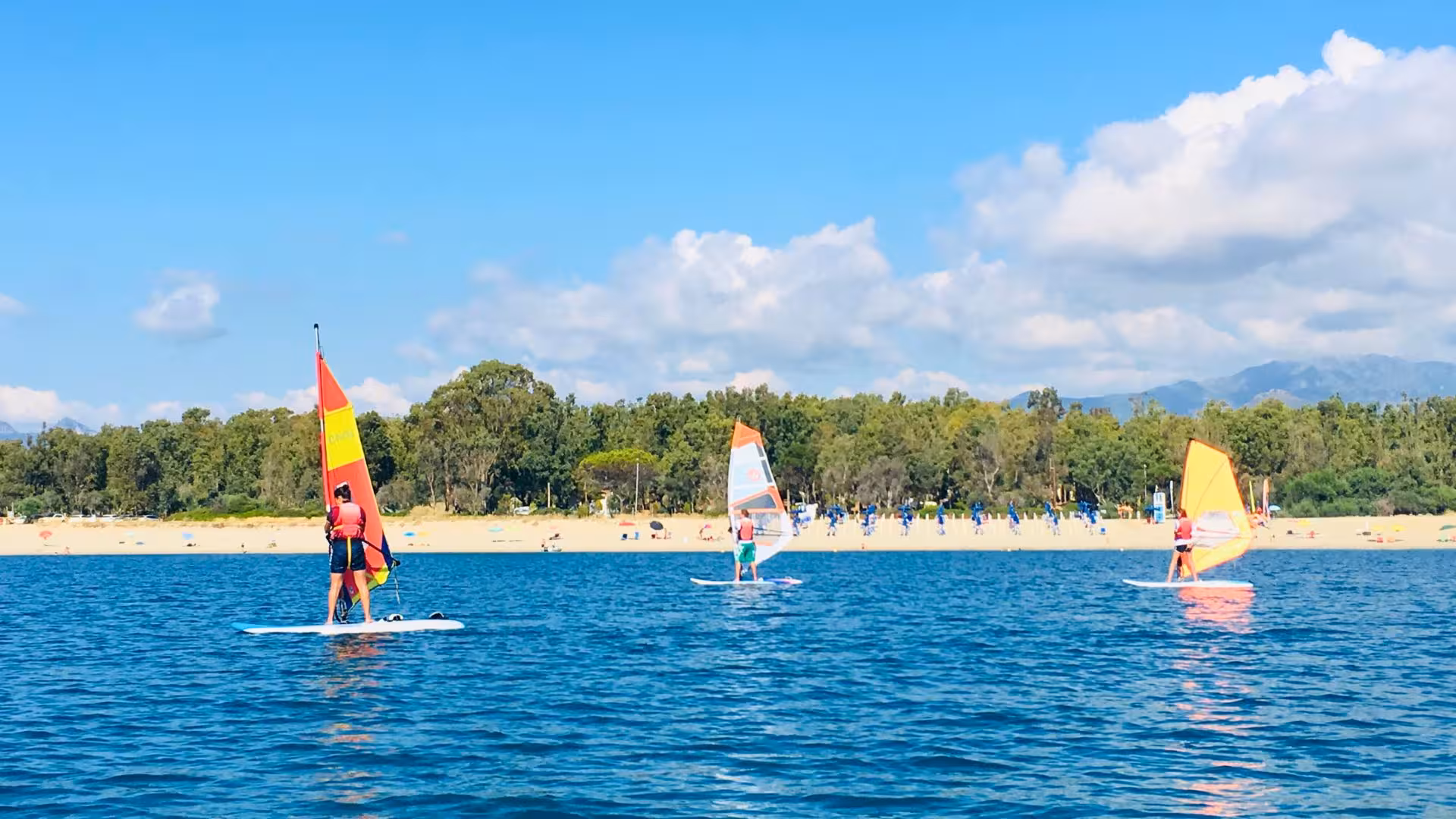 Windsurfers enjoying a sunny day on the calm waters of Bari Sardo, with lush greenery in the background.
