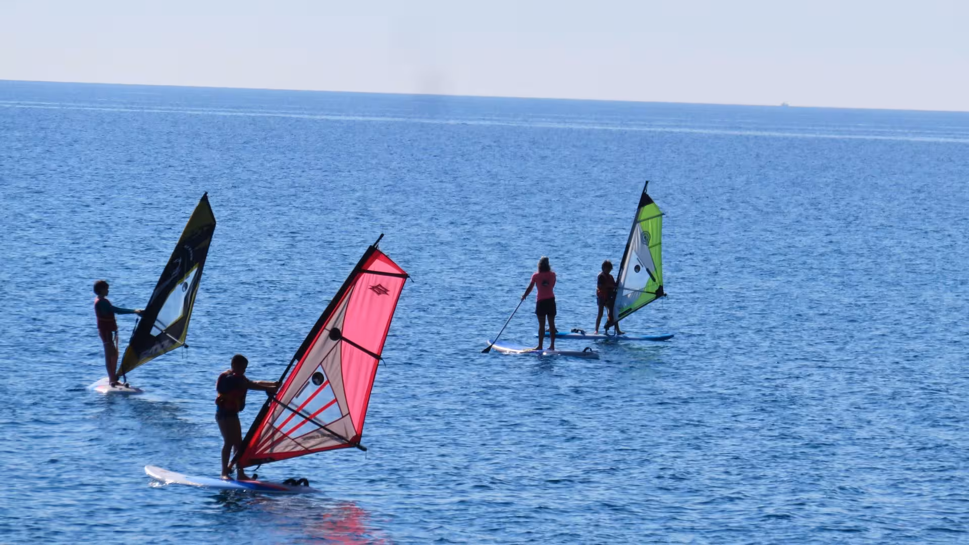 Group of windsurfers gliding over the sparkling blue waters of Bari Sardo during a basic windsurfing course.