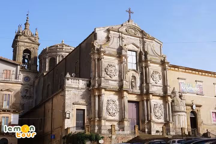 Baroque church façade in Piazza Armerina, featuring intricate stone carvings and a bell tower under a clear blue sky.
