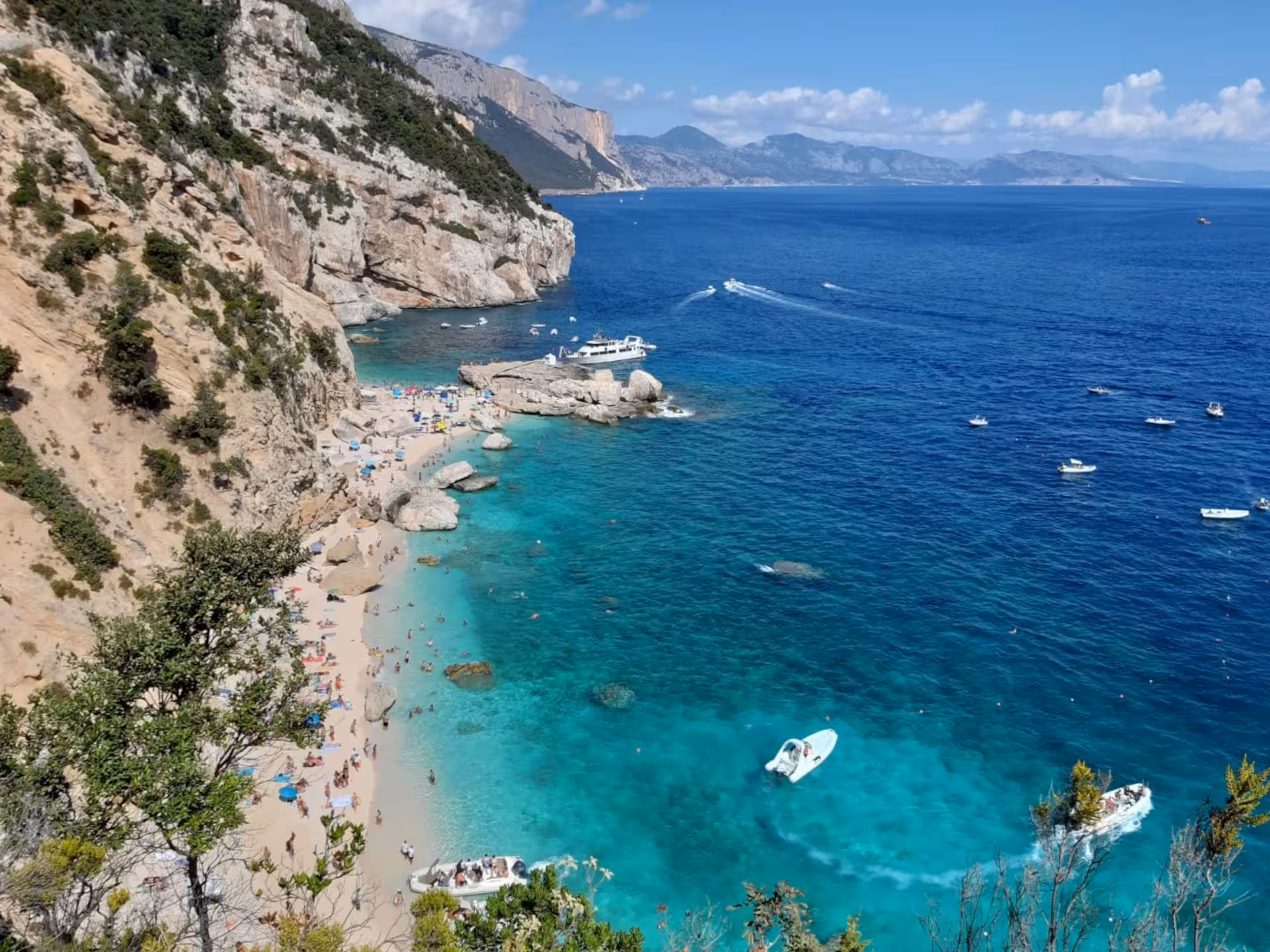 Stunning Cala Mariolu beach with turquoise waters and boats, perfect for a scenic trek from Baunei's coastal cliffs.