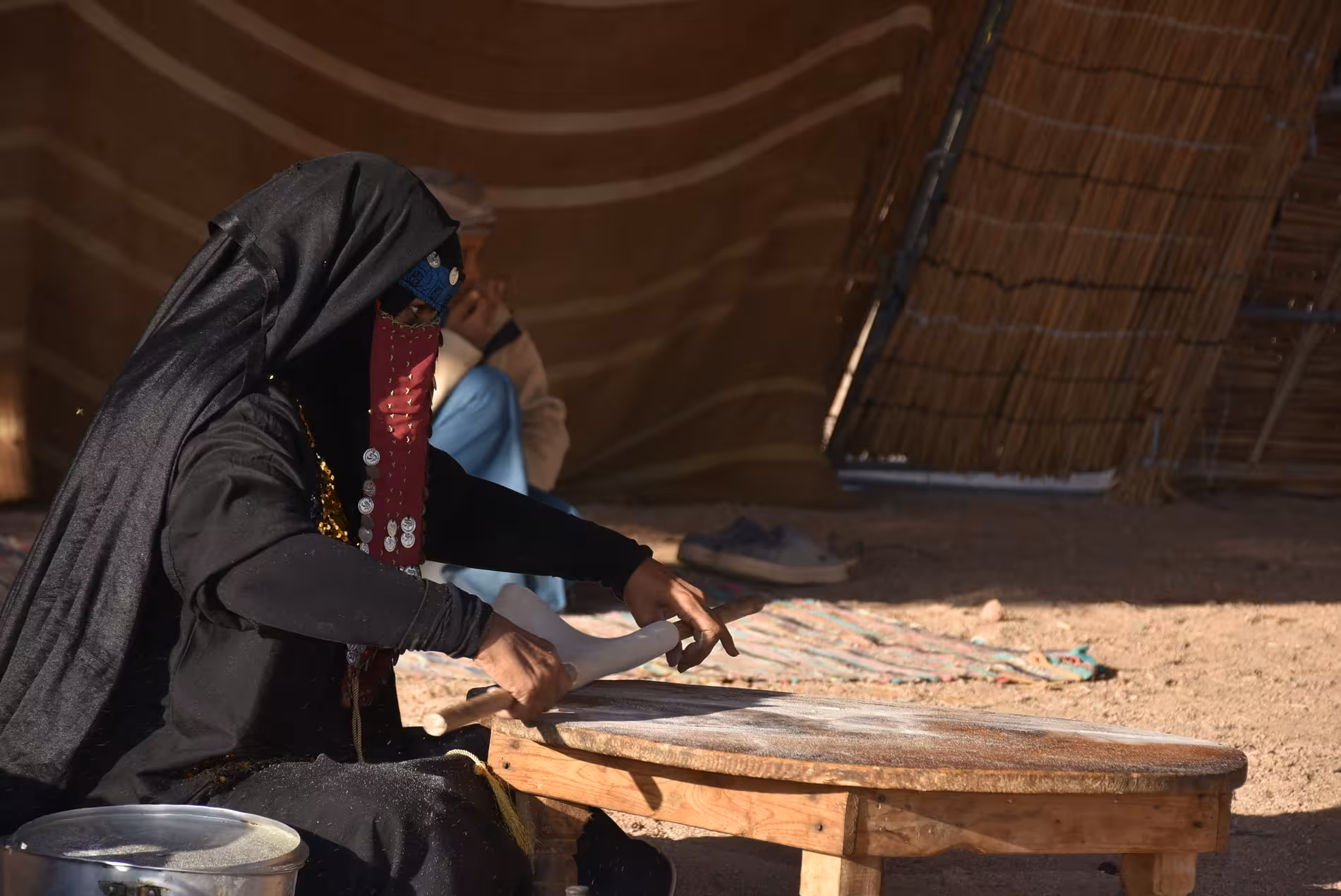 Bedouin woman in traditional black abaya rolling flatbread dough on wooden table at desert camp
